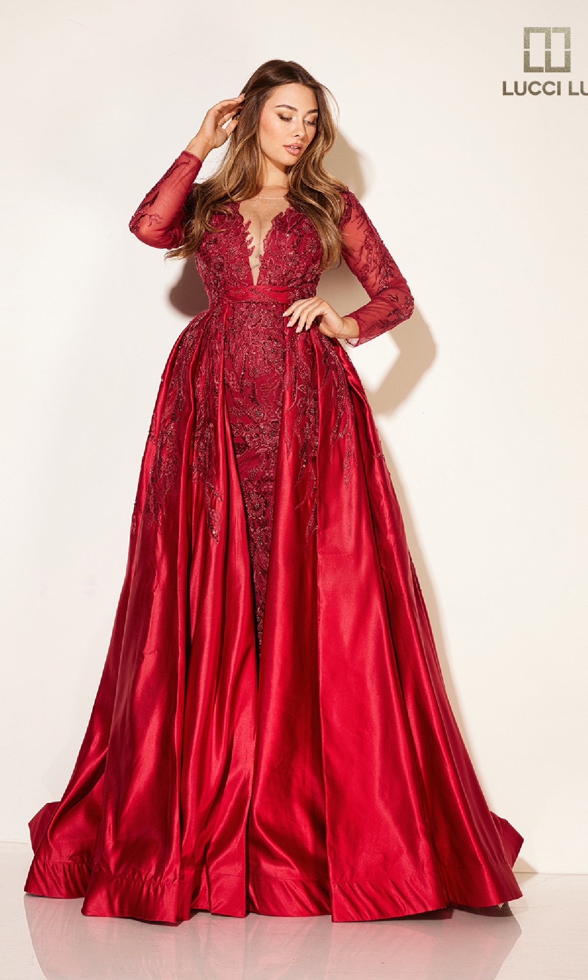 A woman wears the Lucci Lu C8045 long formal dress, featuring long sleeves, deep red color, lace details, a plunging v-neckline, and a full satin skirt, standing against a plain light background.